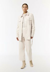 Jasno beżowa kurtka koszulowa z sztucznej skóry z dwiema kieszeniami na piersi, zestawiona z dopasowanymi spodniami o wysokim stanie i kremowymi botkami za kostkę. Gładka tekstura.