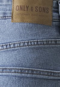 Lys blå denimstoff med synlig søm. En rektangulær brun lærpatch er festet, preget med "ONLY & SONS" og "PASSIONATE JEANS MAKERS."
