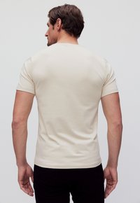 Beige t-shirt met korte mouwen gemaakt van een katoenmix, met een ronde hals en een gladde textuur. Draagt een zwarte broek en is van achteren te zien.
