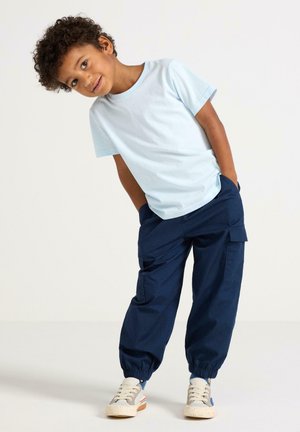 Chlapec se naklání do strany, má na sobě světle modré tričko, tmavě modré kalhoty s kapsami a béžové tenisky, stojí před bílým jednobarevným pozadím.