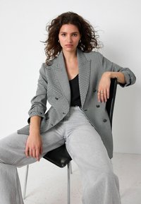Grijze houndstooth dubbelknopen blazer met geklede revers, gecombineerd met lichtgrijze denim broek. Het model zit op een zwarte stoel.
