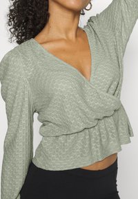 Top texturizado verde claro com decote em V, mangas longas e cintura franzida. Apresenta um design peplum e um padrão geométrico subtil.