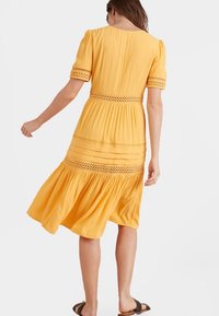 Vestido amarillo con mangas cortas abullonadas, detalles de crochet en la cintura, dobladillo acampanado y tejido suave, combinado con sandalias planas.