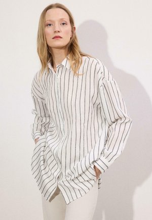 Femme aux cheveux blonds portant une chemise blanche ample à rayures verticales noires et un pantalon blanc, debout les mains dans les poches contre un mur uni.