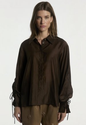 Woman with light brown hair wears loose dark brown long-sleeve blouse with button front and tie details on sleeves, paired with embroidered pants.