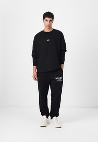Black cotton sweatshirt with ribbed cuffs and a small logo, paired with matching sweatpants featuring a logo and white text accents. White sneakers.
