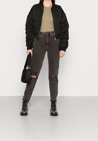 Giubbotto bomber nero con polsini a coste, t-shirt verde oliva, jeans grigio scuro strappati e stivali neri con lacci e punta rotonda. Borsa a spalla nera.