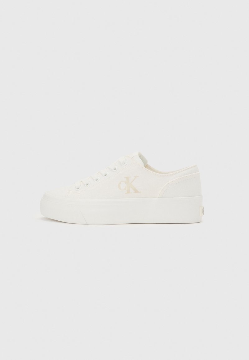 Witte lage sneaker met dikke zool en zijkant "cK" logo, veters aan de voorkant en minimalistisch ontwerp op een neutrale achtergrond.