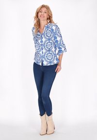 Blusa bianca con motivo floreale blu, maniche a tre quarti e design peplum, abbinata a jeans scuri e stivali da cowboy beige.