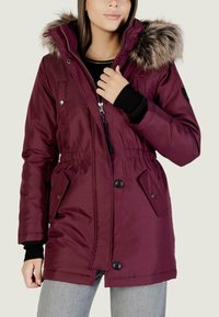 Giacca parka bordeaux con cappuccio in pelliccia sintetica, vita regolabile con coulisse e chiusura lampo davanti. Presenta tasche con bottoni e polsini a coste. Texture liscia.