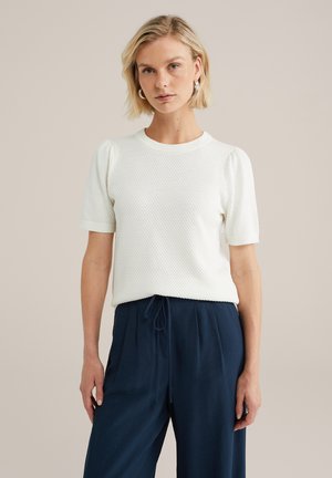 Haut blanc à manches courtes avec un motif en tricot texturé et un col côtelé, associé à un pantalon bleu marine à cordon de serrage, mettant en valeur une coupe décontractée.