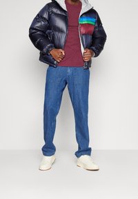 Doudoune bleu marine avec capuche, ornée d'un patch multicolore sur la poitrine. Chemise bordeaux, jean bleu et baskets blanches complètent la tenue.