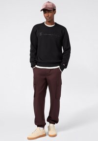 Felpa nera con logo "Champion", pantaloni cargo bordeaux con tasche e sneaker beige. Gli accessori includono un cappellino rosa.