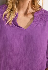 Un haut violet texturé avec un décolleté en V et des détails de fronces près du col, des manches courtes et une coupe décontractée.