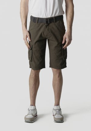 Mand iført olivengrønne cargoshorts med bælte, hvide sneakers og hvid t-shirt, stående mod en ensartet, lys grå baggrund.