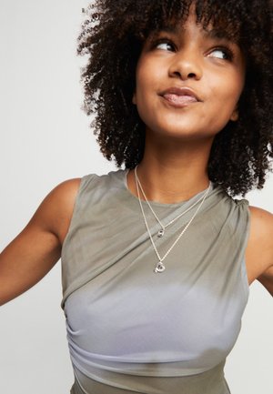 Femme aux cheveux bouclés portant un haut sans manches avec un dégradé de gris à violet clair et des colliers argentés superposés, regardant légèrement vers le haut et sur le côté.