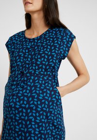 Niebieska sukienka ciążowa w wzory z rękawami na kapelusz, okrągłym dekoltem, elastycznym paskiem z możliwością związania oraz kieszeniami bocznymi.