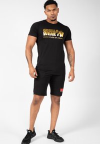 Fekete pamut póló arany "GORILLA WEAR UNITED STATES OF AMERICA" felirattal, fekete rövidnadrággal, amelyen piros logó található, fekete sportcipővel viselve.