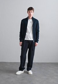 Temno modra jakna na zadrgo čez belo majico, kombinirana z ohlapnimi črnimi cargo hlačami in belimi športnimi čevlji. Minimalističen dizajn s čistimi linijami.