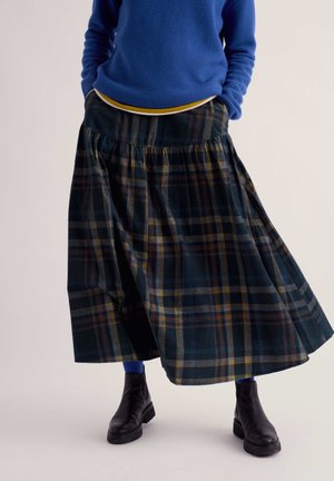 Person wearing a blue sweater, dark plaid long skirt, blue leggings, and black ankle boots, standing with hands in pockets.