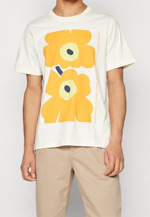 Mann trägt weißes T-Shirt mit großem, leuchtend gelbem Blumenmotiv und beiger Hose vor einfarbigem Hintergrund.