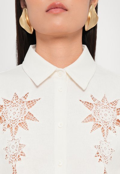 White shirt with a collar, featuring intricate lace cutouts in star patterns. Gold, sculptural earrings are visible. Smooth texture.