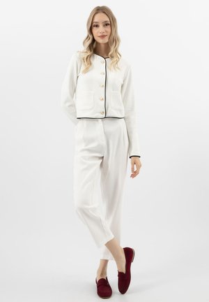 Junge Frau steht mit entspannter Haltung, trägt eine weiße Jacke mit Knöpfen, passende weiße Hose und bordeauxrote Loafers vor einfachem Hintergrund.