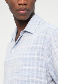 Εγγύς λήψη ενός ατόμου που φοράει ανοιχτό μπλε πουκάμισο με κοντά μανίκια, διακριτικές οριζόντιες ρίγες και γιακά, σε λευκό φόντο.