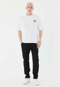 Hvid bomulds t-shirt med et lille sort logo på brystet, parret med sorte bukser og hvide sneakers. Enkel, afslappet påklædning.