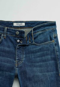 Blauwe slim-fit Salsa Jeans met gedeeltelijk dichtgeknoopte knoopsluiting, zichtbare stiknaden en voorzakken op een neutrale achtergrond.