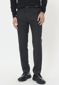 Férfi sötétszürke kockás öltönynadrágban, fekete övvel, fekete hosszú ujjú ingben és fényes fekete bőrcipőben áll egy fehér háttér előtt.