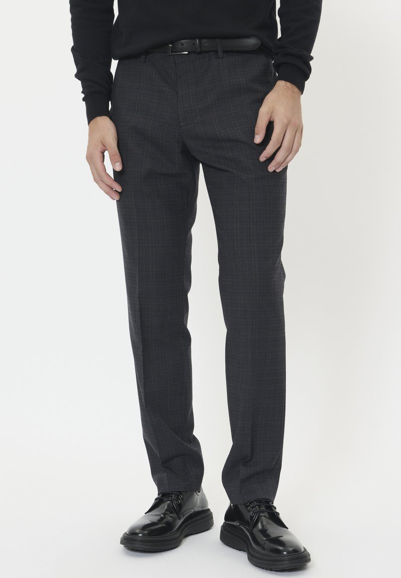 Férfi sötétszürke kockás öltönynadrágban, fekete övvel, fekete hosszú ujjú ingben és fényes fekete bőrcipőben áll egy fehér háttér előtt.