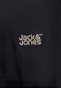 Κέντημα του ονόματος της μάρκας "Jack & Jones" με μπεζ νήμα σε μαύρο ύφασμα, κεντραρισμένο και καθαρά ορατό.