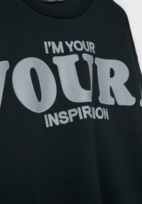 Čierna mikina s okrúhlym výstrihom, na ktorej sa nachádzajú veľké, tučné biele písmená, ktoré čítajú "JA SOM TVOJ" nad "INŠPIRÁCIA" v menšom písme.