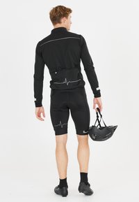 Tenue de cyclisme noire comprenant un haut à manches longues avec des accents réfléchissants et un short au motif de battements de cœur. Un casque noir est tenu à la main.