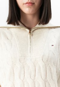 Femme portant un pull crème en maille torsadée avec une fermeture éclair dorée à demi-zip et un col côtelé, arborant un petit logo rouge, blanc et bleu sur la poitrine.