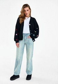 Žena s dlhými vlasmi, ktorá má na sebe čierny kardigán s farebnými hviezdami, biely top, svetlomodré džínsy a čierne platformové topánky.