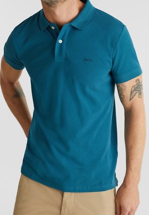 Mężczyzna noszący turkusową koszulkę polo z krótkim rękawem z dwoma białymi guzikami oraz beżowe spodnie, pokazujący tatuaże na obu ramionach.