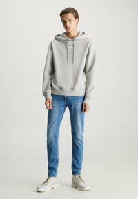 Sudadera gris claro con cordones, combinada con jeans azules y botas blancas con cordones. La sudadera tiene un logo en el frente y puños acanalados.