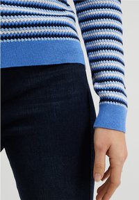 Blå stickad tröja med ett strukturerat mönster som har diamantskikt i marinblått, ljusblått och vita ränder, i kombination med mörka jeans.