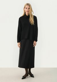 Mustreeritud must sall-sweater, millel on kaelus, kombineeritud pika musta seeliku, hõõguvate mustade sukkade ja mustade terava nina sandaalidega, neutraalse tausta taustal.