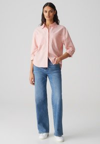 Helles rosa Hemd mit zwei Brusttaschen, hochgekrempelten Ärmeln, kombiniert mit hochgeschnittenen blauen Jeans und weißen Turnschuhen.