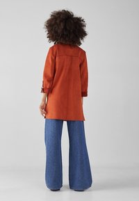 Abrigo de ante color óxido con mangas de tres cuartos, corte recto y detalle de yugo en la parte posterior, combinado con jeans de mezclilla azul de pierna ancha.