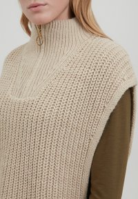 Beige gestrickter ärmelloser Pullover mit hohem Kragen und Reißverschluss. Verfügt über ein strukturiertes Muster und ein geschichtetes Design über einem braunen Oberteil.