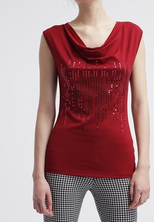 Mujer con top rojo sin mangas con escote drapeado y patrón de lentejuelas vertical, combinado con pantalones de pata de gallo en blanco y negro.