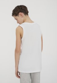 Camicia bianca senza maniche realizzata in morbido cotone. Texture liscia, design semplice, con scollatura a girocollo e taglio dritto. Indossata con pantaloni grigi.