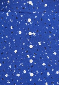 Tessuto floreale blu con fiori bianchi e neri, piccoli punti bianchi. Presenta bottoni verticali al centro, superficie liscia.