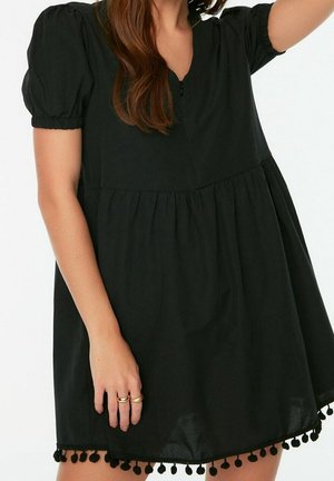 Kobieta w czarnej sukience z bufkami i ozdobą pomponów na dole, pokazująca dłoń z dwoma złotymi pierścionkami.