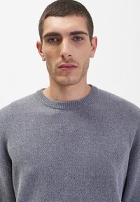 Grijze sweatervest met een gestructureerde stof, voorzien van een ribbelachtige halslijn en een ontspannen pasvorm. Het materiaal lijkt zacht en warm.