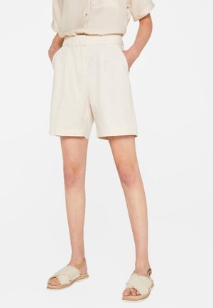 Person iført højtaljede, cremefarvede shorts med forreste folder og lommer, kombineret med cremefarvede sandaler med krydsede stropper.